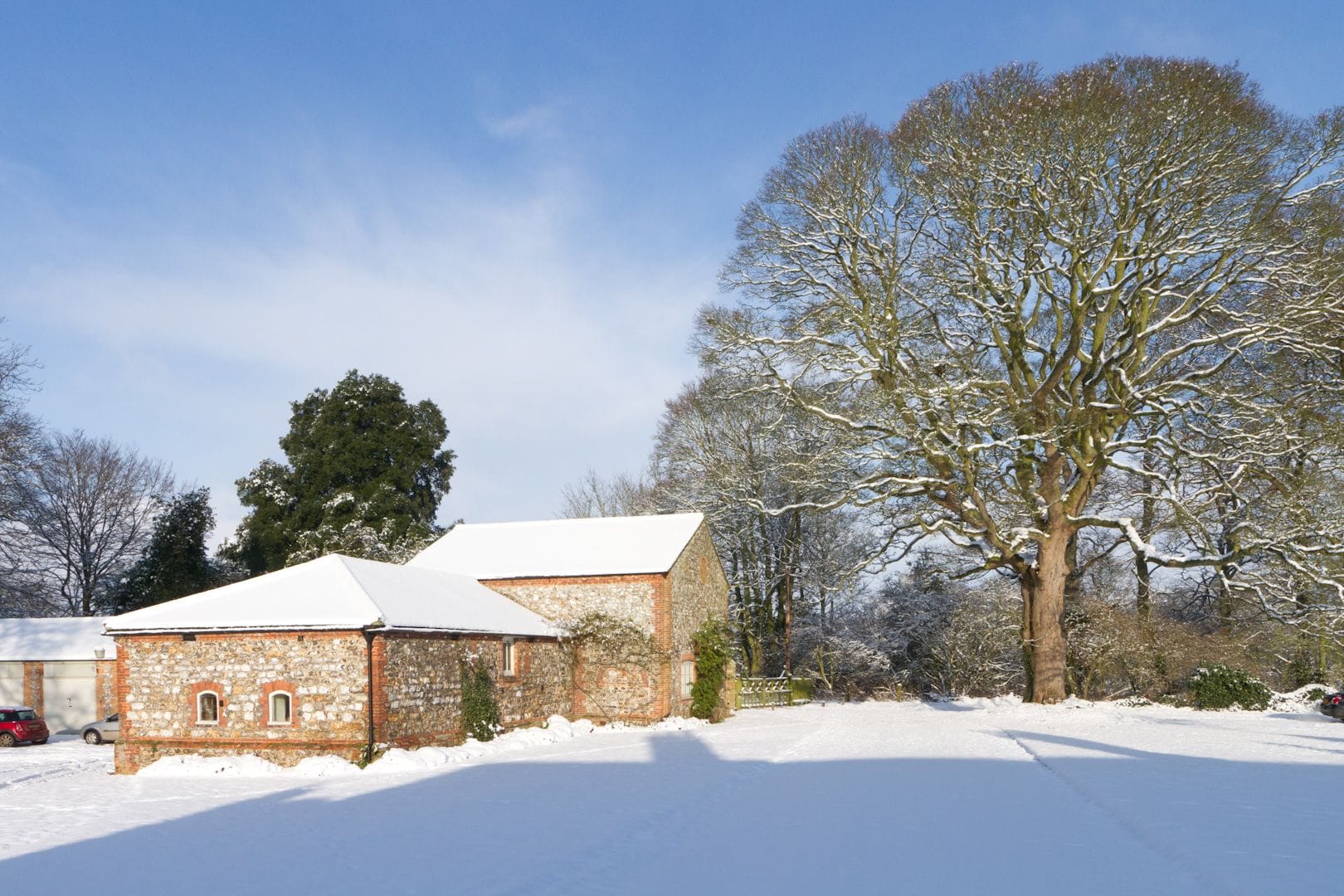Snow-covered flint outbuildings and trees at Anmer Hall under a blue sky
