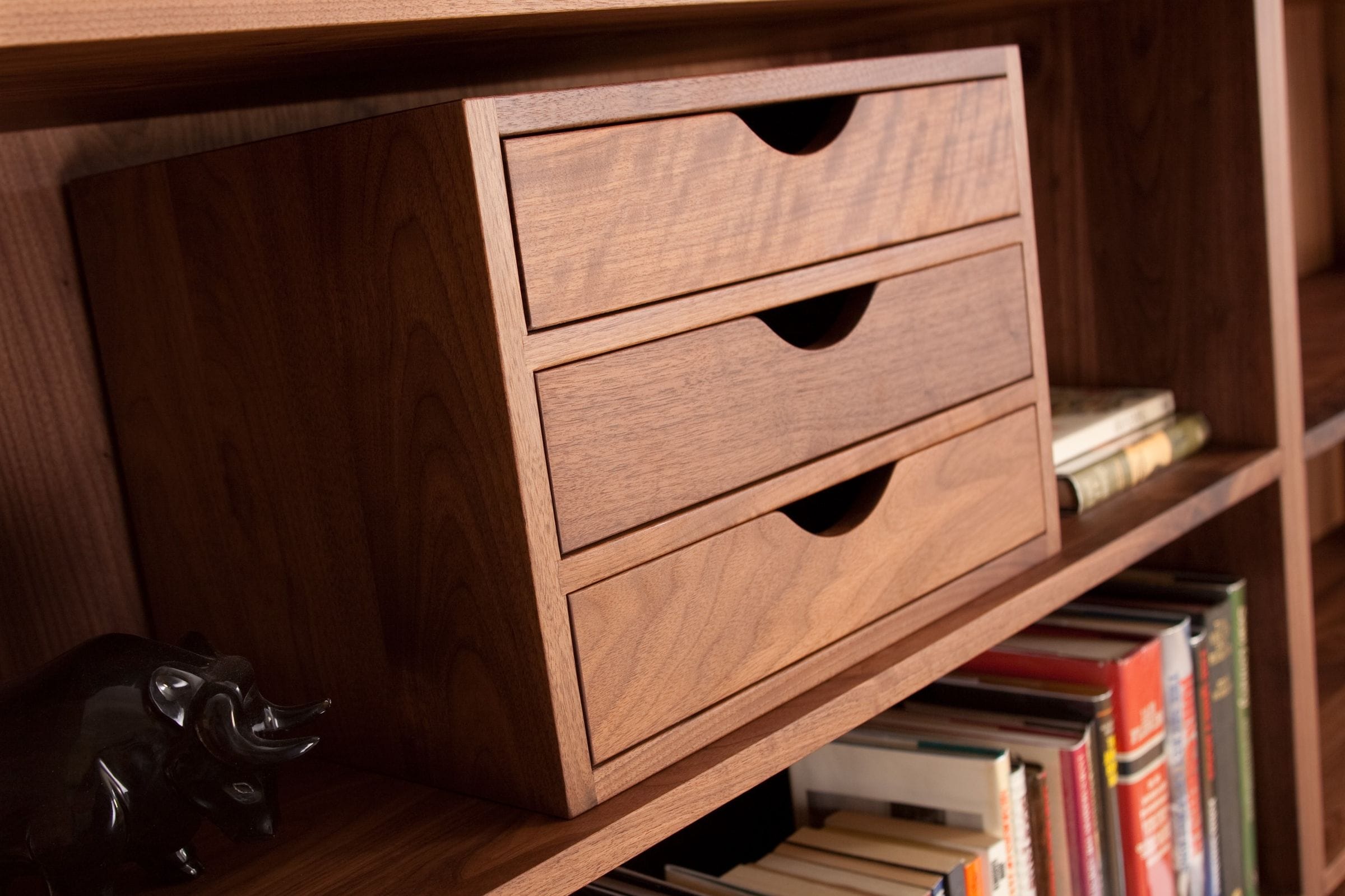 Three-drawer walnut filing box sitting on a bookshelf beside rows of books