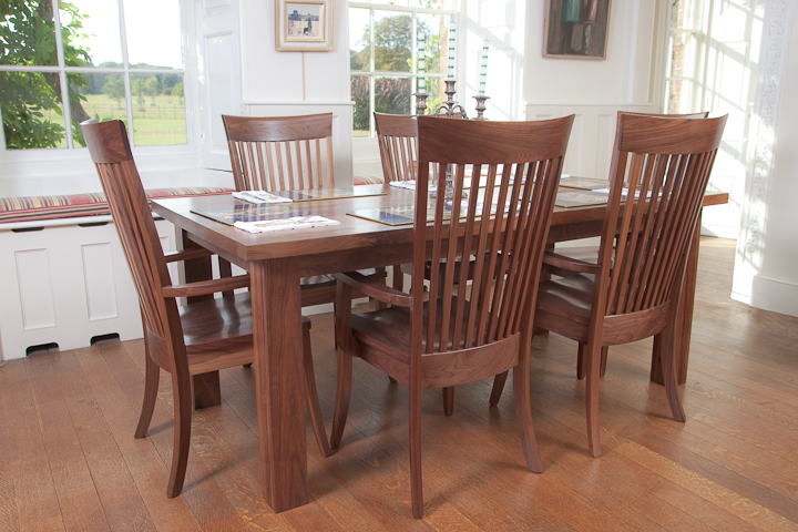 Walnut dining table and six slat-back chairs beside a bright window seat